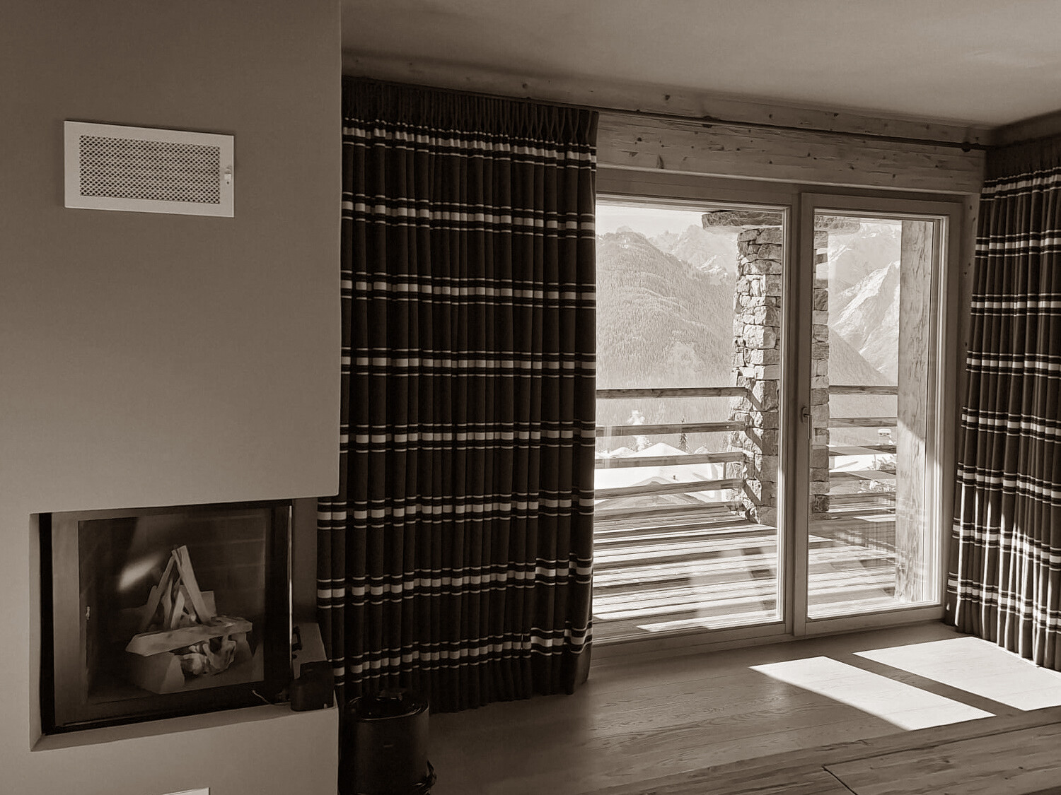 A cozy alpine bedroom featuring a built-in fireplace, rustic wood details, and deep-striped curtains framing breathtaking mountain views through a large glass door.