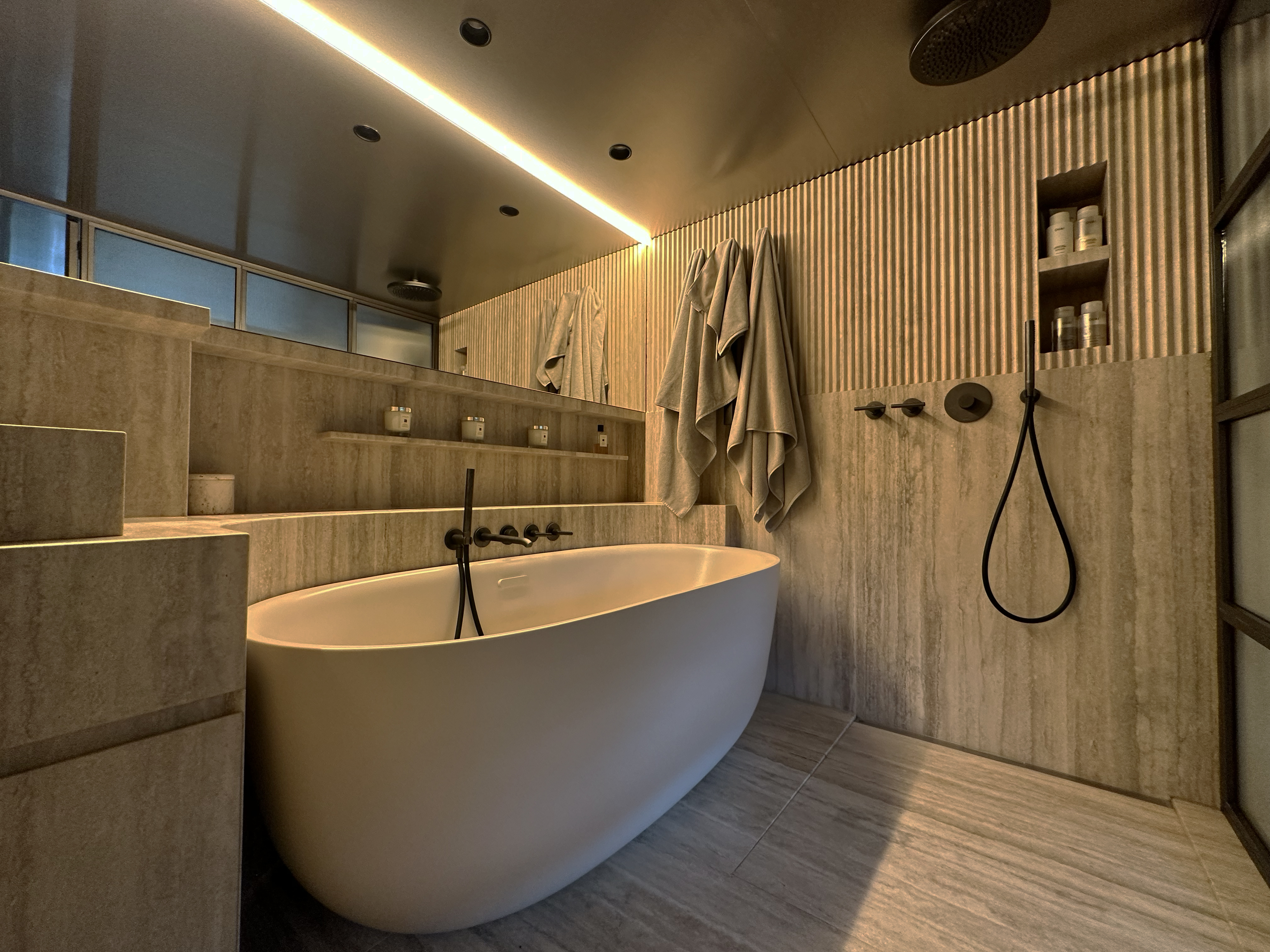 Luxurious spa-style bathroom featuring a freestanding tub nestled against warm stone steps, textured wall paneling, and recessed shelving. Matte black fixtures and soft ambient lighting complete the serene and refined atmosphere, offering a true retreat for relaxation. Luxurious spa-style bathroom featuring a freestanding tub nestled against warm stone steps, textured wall paneling, and recessed shelving. Matte black fixtures and soft ambient lighting complete the serene and refined atmosphere, offering a true retreat for relaxation.