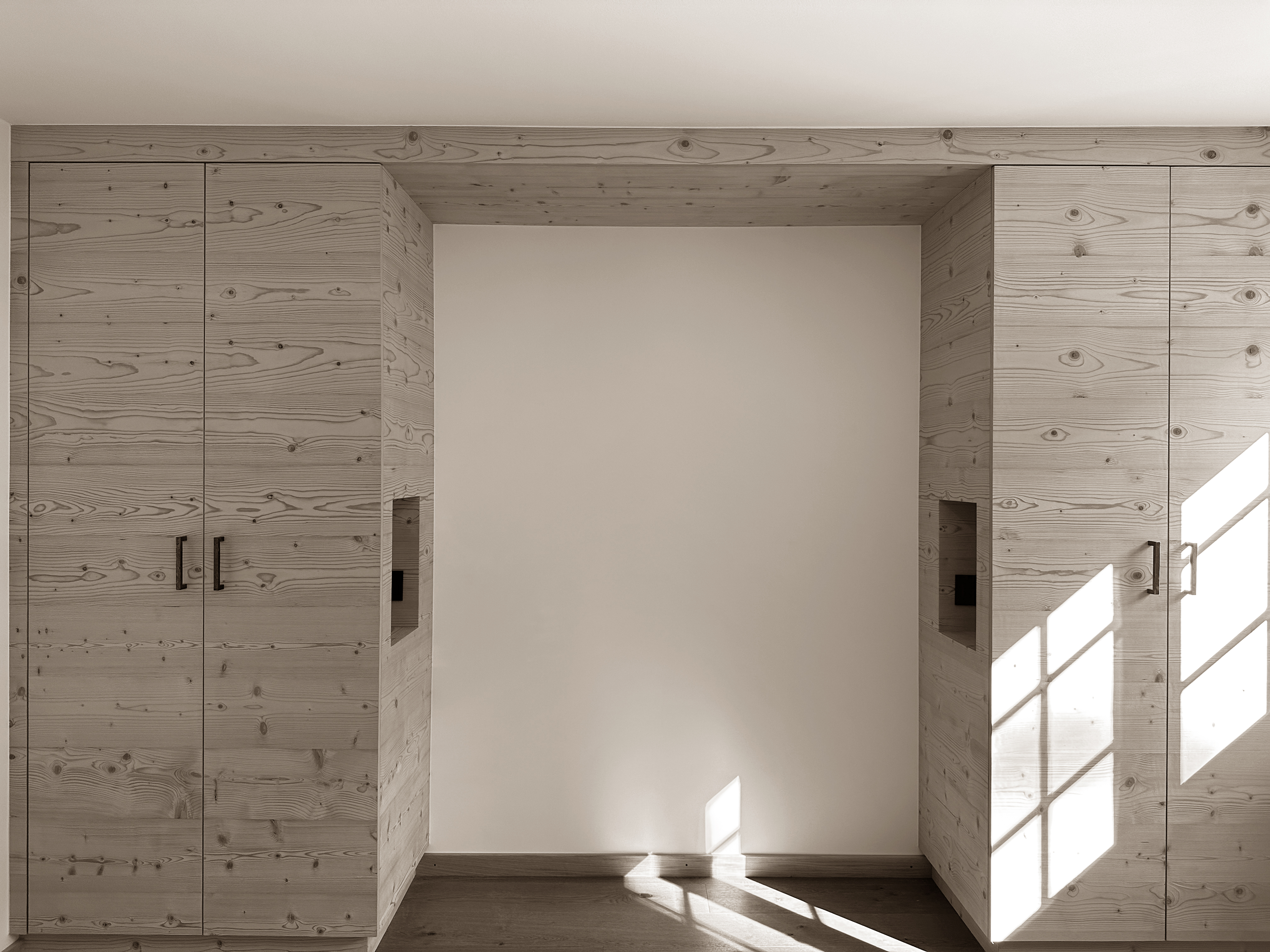 Refined alpine interior with built-in wooden wardrobes framing an open alcove designed to accommodate a bed, bathed in natural light that enhances the clean lines, warm tones, and serene atmosphere of the space.
