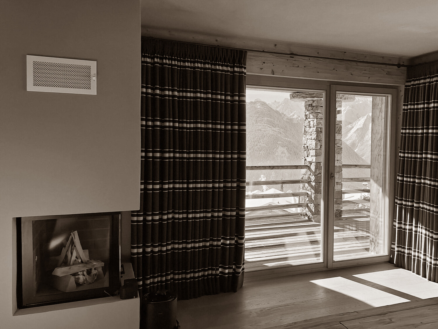 A cozy alpine bedroom featuring a built-in fireplace, rustic wood details, and deep-striped curtains framing breathtaking mountain views through a large glass door.