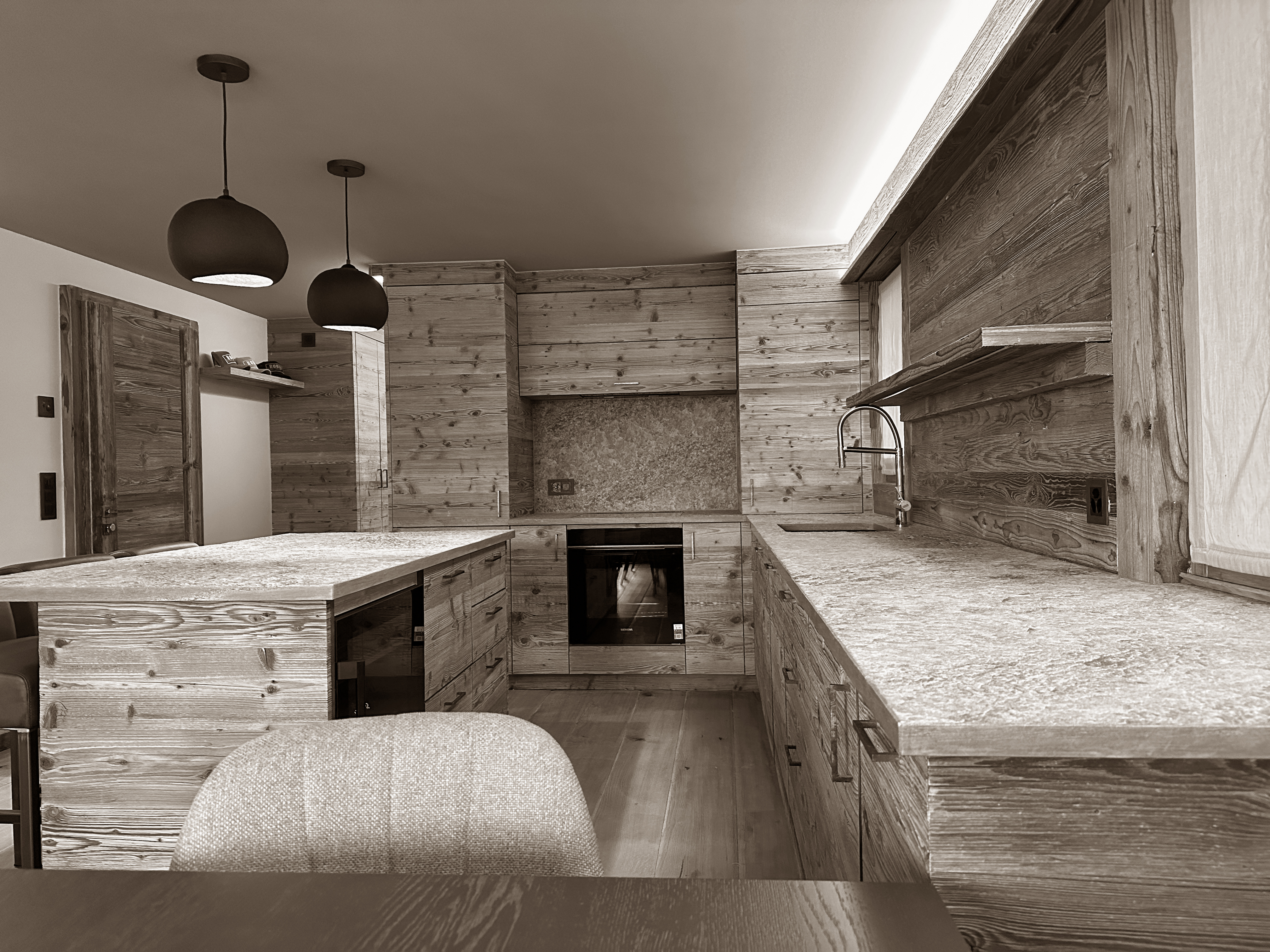 Expansive open kitchen with handcrafted wooden cabinetry and stone countertops, blending rustic charm with modern design. Pendant lighting adds a contemporary touch, while the spacious layout invites convivial cooking and shared alpine moments.